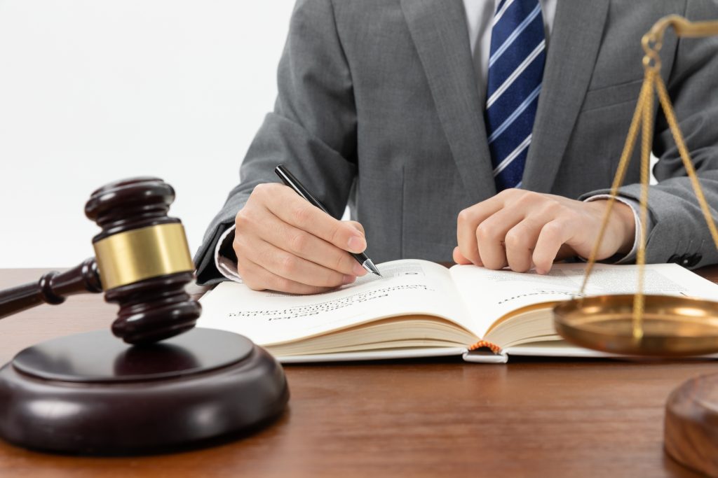 Homem caucasiano de meia-idade, vestindo um terno cinza e uma gravata azul, sentado em uma mesa, escrevendo em um caderno, com um martelo na mesa. Uma cena que remete à Assessoria Jurídica.