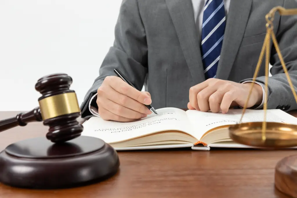 Homem caucasiano de meia-idade, vestindo um terno cinza e uma gravata azul, sentado em uma mesa, escrevendo em um caderno, com um martelo na mesa. Uma cena que remete à Assessoria Jurídica.