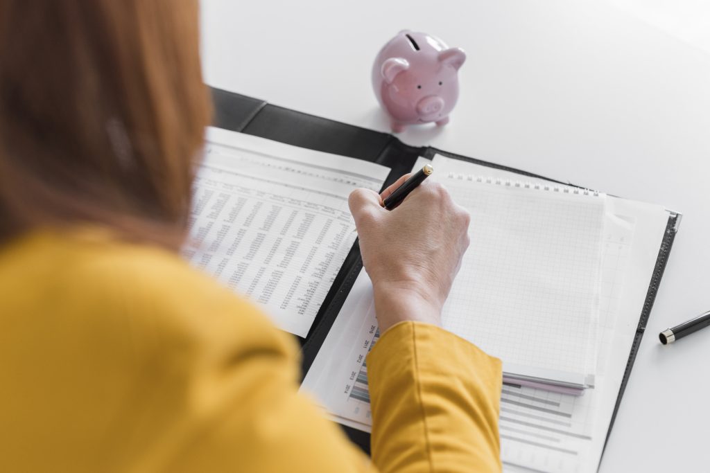 Uma jovem de suéter amarelo sentada em uma mesa, escrevendo em um caderno e olhando para um cofrinho rosa sobre a mesa. Essa cena pode representar a organização financeira para a Recuperação de Créditos Tributários.