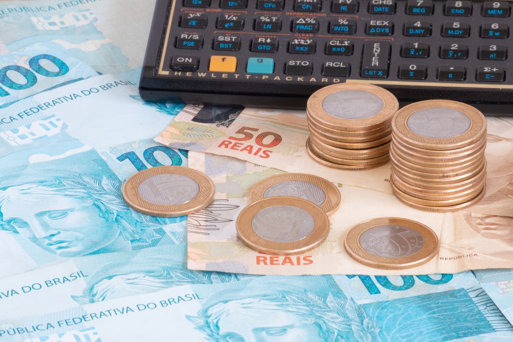 Pilhas de moedas e notas de real brasileiro em uma calculadora, representando conceitos financeiros como contabilidade, orçamento ou câmbio, o que inclui o cálculo de PIS e COFINS.