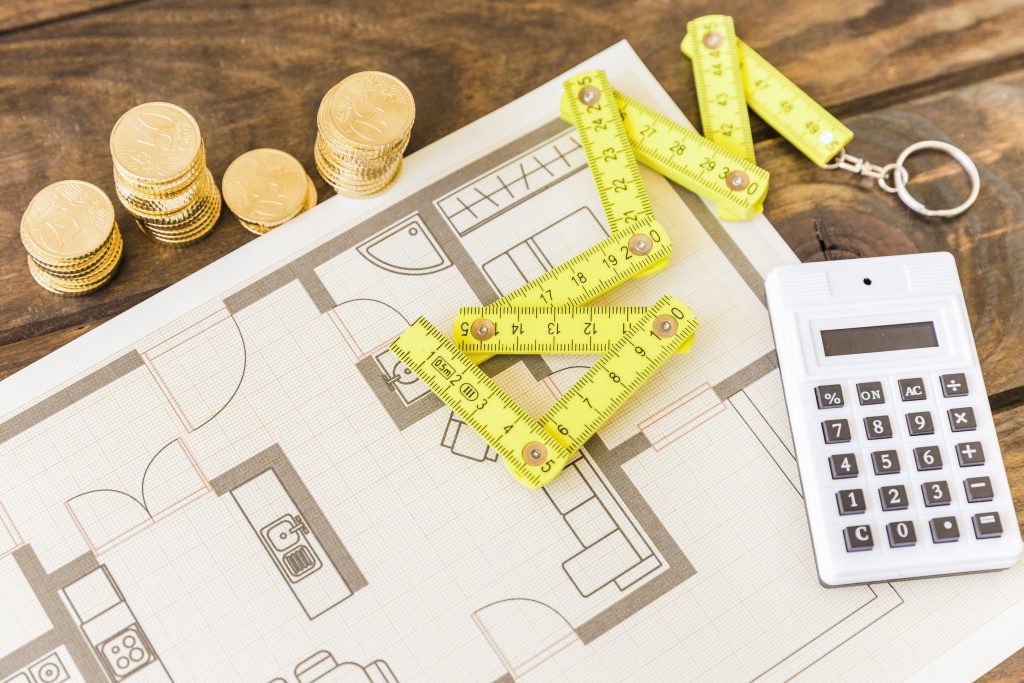 Um plano de construção com uma fita métrica amarela, moedas e uma calculadora sobre uma mesa de madeira, todos essenciais para o Pagamento de taxas.