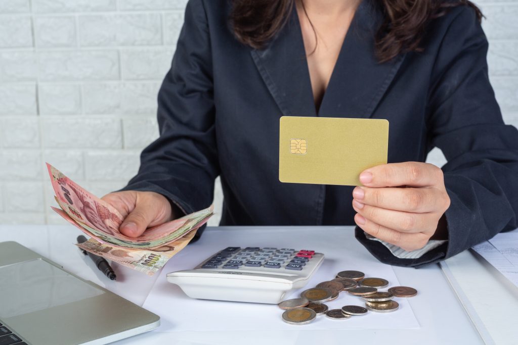 Uma mulher de meia-idade, de terno preto, segura um cartão de crédito e conta dinheiro em uma mesa com calculadora e moedas. Essa cena reflete a gestão financeira em casos de Restituições e compensações tributárias.