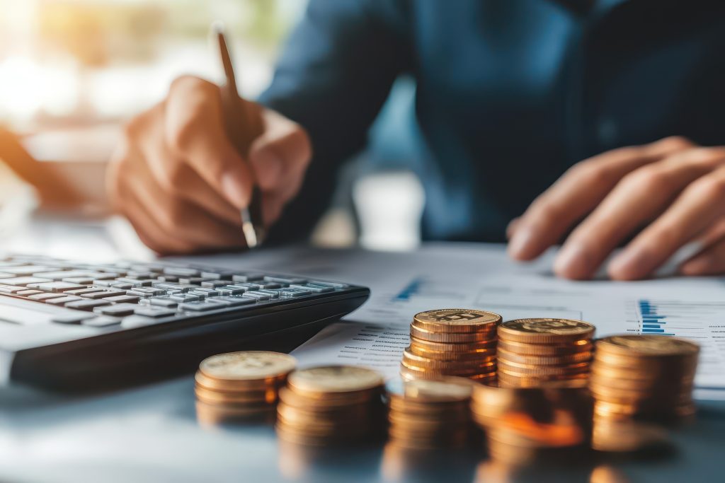 As mãos de uma pessoa usando uma calculadora e contando pilhas de moedas em uma mesa. Cena que ilustra a auditoria para garantir o Compliance fiscal.