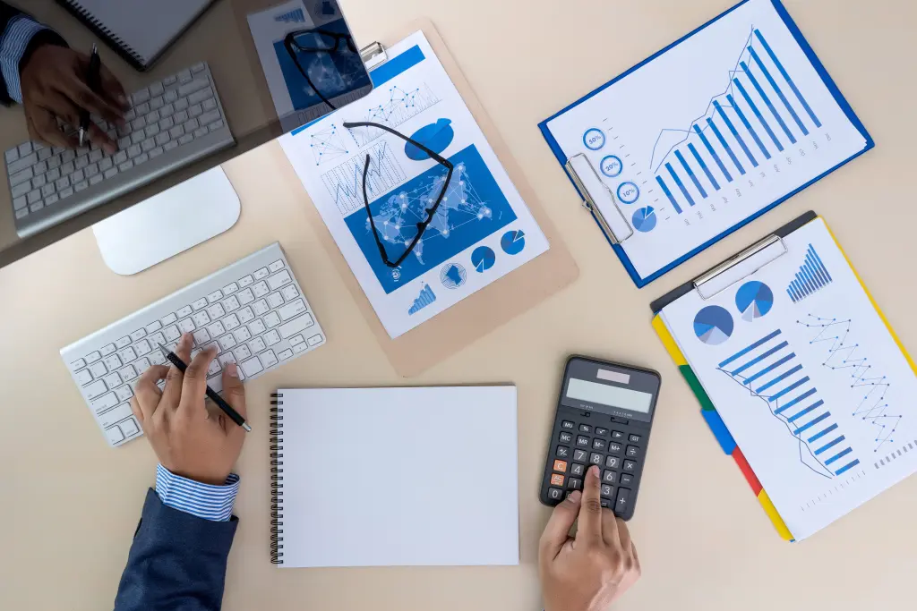 Mãos trabalhando em documentos financeiros, com gráficos e calculadora. Cena ilustra a aplicação de Estratégias de otimização tributária.