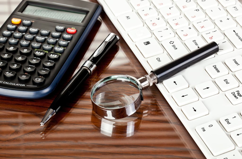 Uma visão de perto de uma mesa com uma calculadora, uma caneta preta, uma lupa e um teclado branco. A superfície é de madeira, refletindo os itens colocados nela
