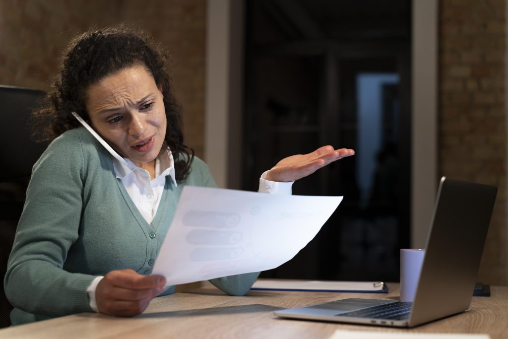 Mulher frustrada ao telefone, segurando um papel com laptop à frente. A cena sugere problemas de Pagamento Indevido de Impostos.