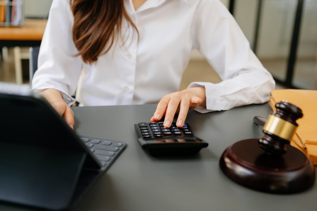 Mãos de uma mulher usando calculadora em uma mesa com um martelo de juiz. Cena que pode envolver o cálculo e o Pagamento de imposto, seguindo as regulamentações fiscais.