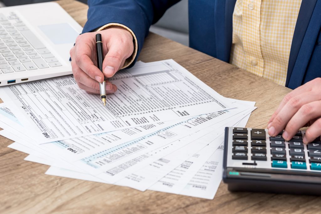 Pessoa preenchendo formulários fiscais com calculadora, em uma mesa de trabalho. Imagem relacionada à complexa Recuperação de tributos.
