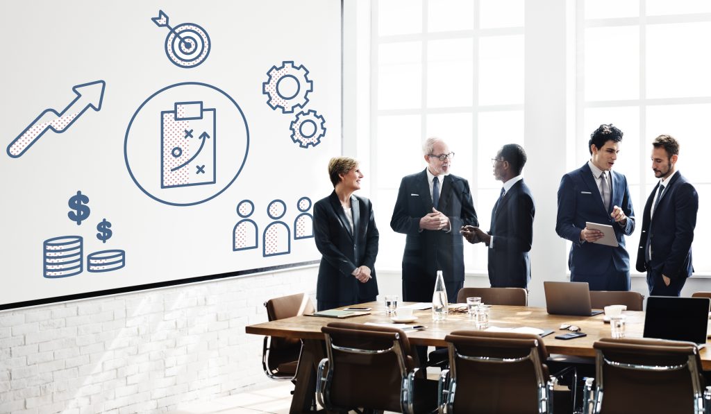 Grupo de profissionais de negócios reunidos em uma sala de reunião moderna, com um quadro branco exibindo ícones de crescimento, metas, estratégia e equipe.