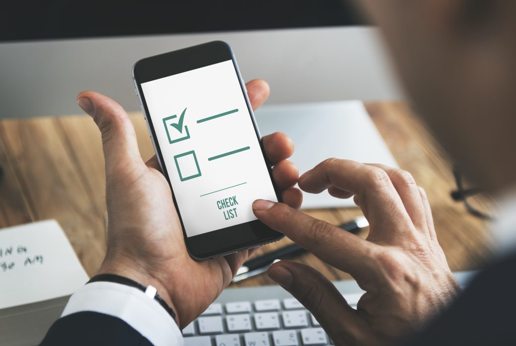 Close-up das mãos de um homem interagindo com um celular que mostra um aplicativo de lista de tarefas. Na tela, há duas caixas de seleção, uma já marcada com um símbolo de "check" verde e a outra ainda vazia, acompanhadas de linhas horizontais. Abaixo, lê-se "CHECK LIST". Este cenário digital remete à necessidade de um checklist jurídico para empresas do varejo para a gestão eficiente de suas obrigações legais.