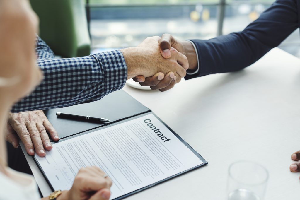 Duas pessoas apertam as mãos sobre uma mesa, com um contrato em primeiro plano, simbolizando o fechamento de um acordo. A cena representa a formalização de Cláusulas contratuais em relações comerciais.