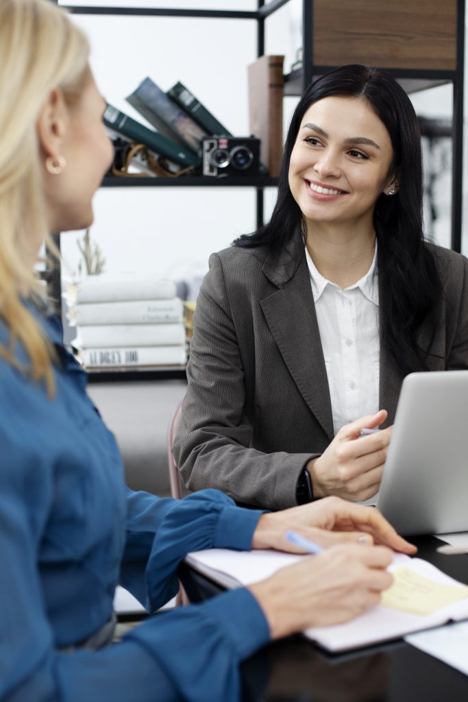 Duas mulheres estão reunidas em um ambiente profissional. Uma delas, com cabelos escuros e vestindo um blazer cinza sobre uma blusa branca, sorri enquanto olha para a outra. A segunda mulher, de costas para a câmera, tem cabelos loiros e veste uma blusa azul. Elas parecem estar envolvidas em uma conversa ou reunião de trabalho, com um laptop aberto e anotações sobre a mesa, talvez discutindo estratégias de jurídico preventivo para a empresa. O ambiente sugere uma atmosfera colaborativa e focada em soluções.