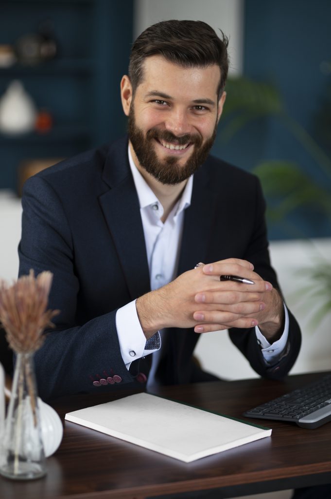 Um homem sorridente, com barba bem cuidada e vestindo um elegante blazer azul sobre uma camisa branca, está sentado à sua mesa de trabalho. Ele segura uma caneta entre as mãos unidas, com um olhar amigável e profissional. À sua frente, repousam um caderno branco e um teclado de computador, sugerindo um ambiente de trabalho focado em organização e estratégia, talvez relacionado a planejamento jurídico preventivo. Um vaso com flores secas adiciona um toque de serenidade ao espaço.