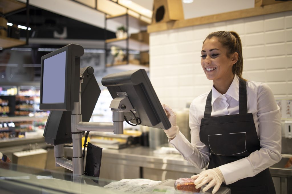 Uma atendente sorridente em um açougue de supermercado, com avental e luvas, prepara um corte de carne. A cena ilustra o ambiente de trabalho que pode levar a Ações Trabalhistas em supermercados se as condições não forem adequadas.