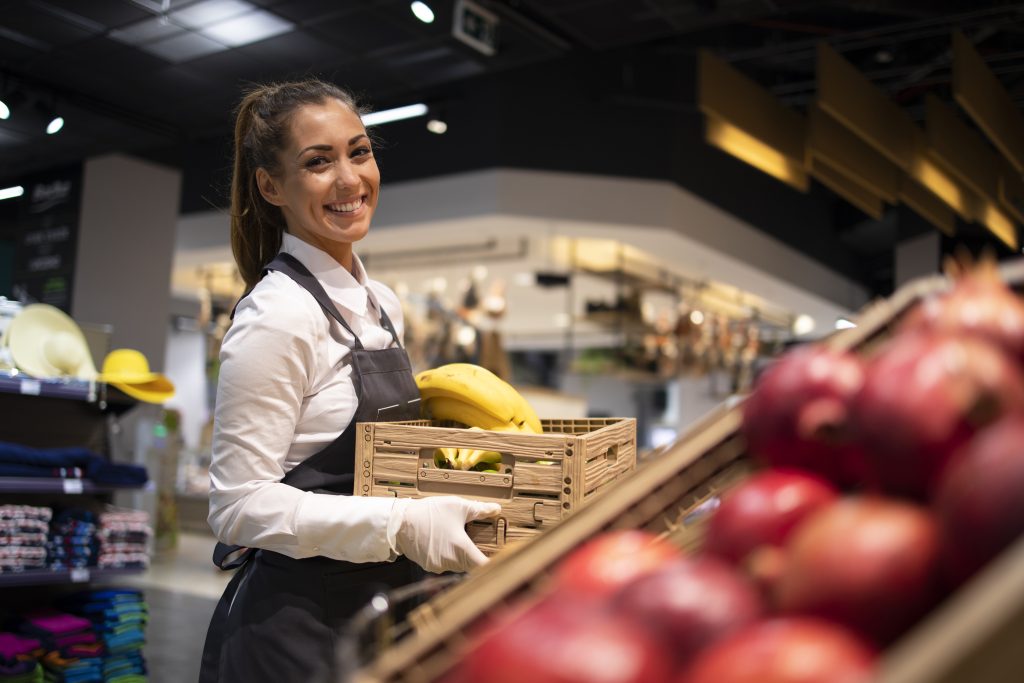 Uma funcionária sorridente de supermercado, usando luvas e avental, reabastece a seção de frutas. A imagem destaca as atividades diárias que podem gerar Ações Trabalhistas em supermercados se os direitos não forem respeitados.