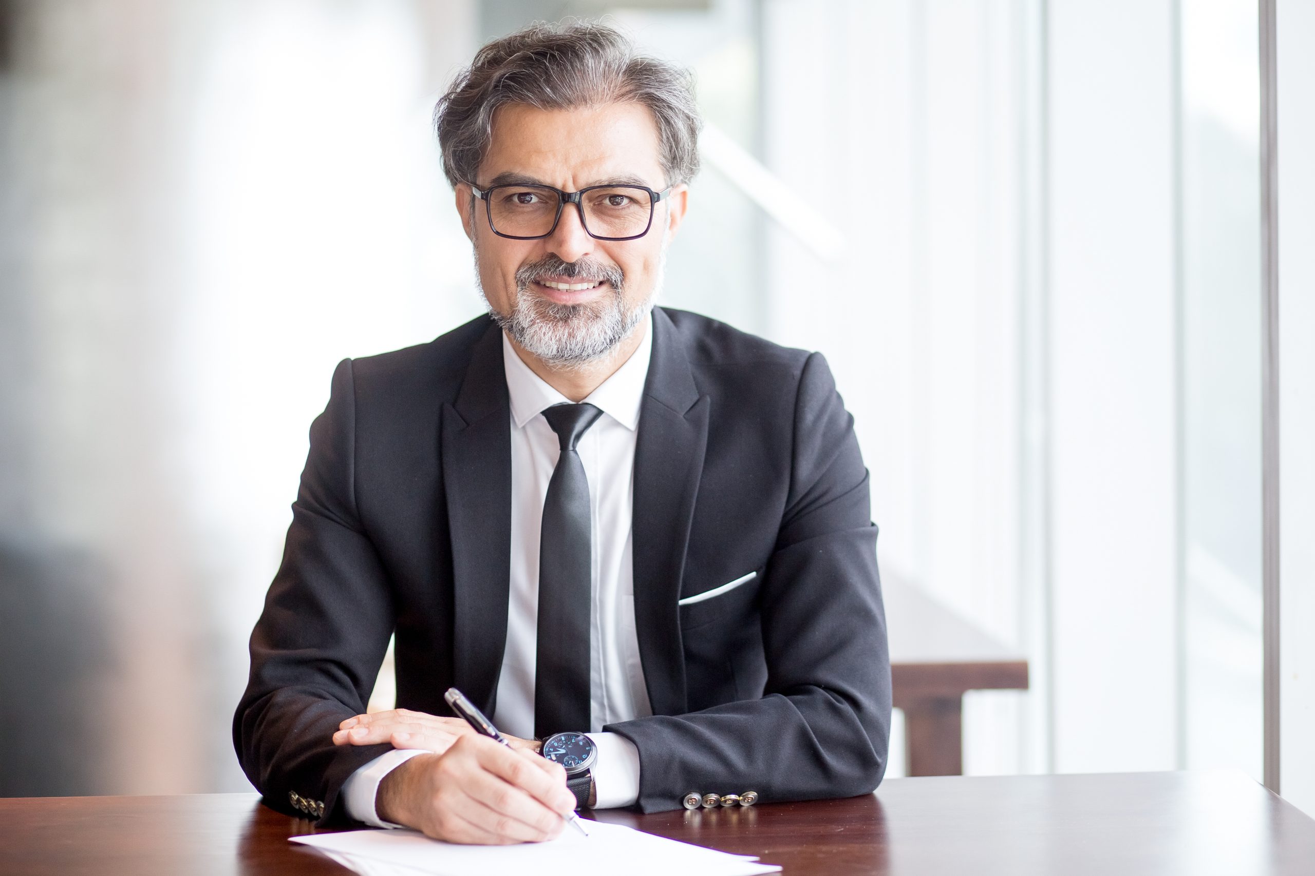 Um homem de meia-idade, com barba e óculos, trajando terno, sorri enquanto escreve. Sua postura reflete a atenção aos detalhes, essencial na Advocacia Preventiva para evitar futuros litígios.