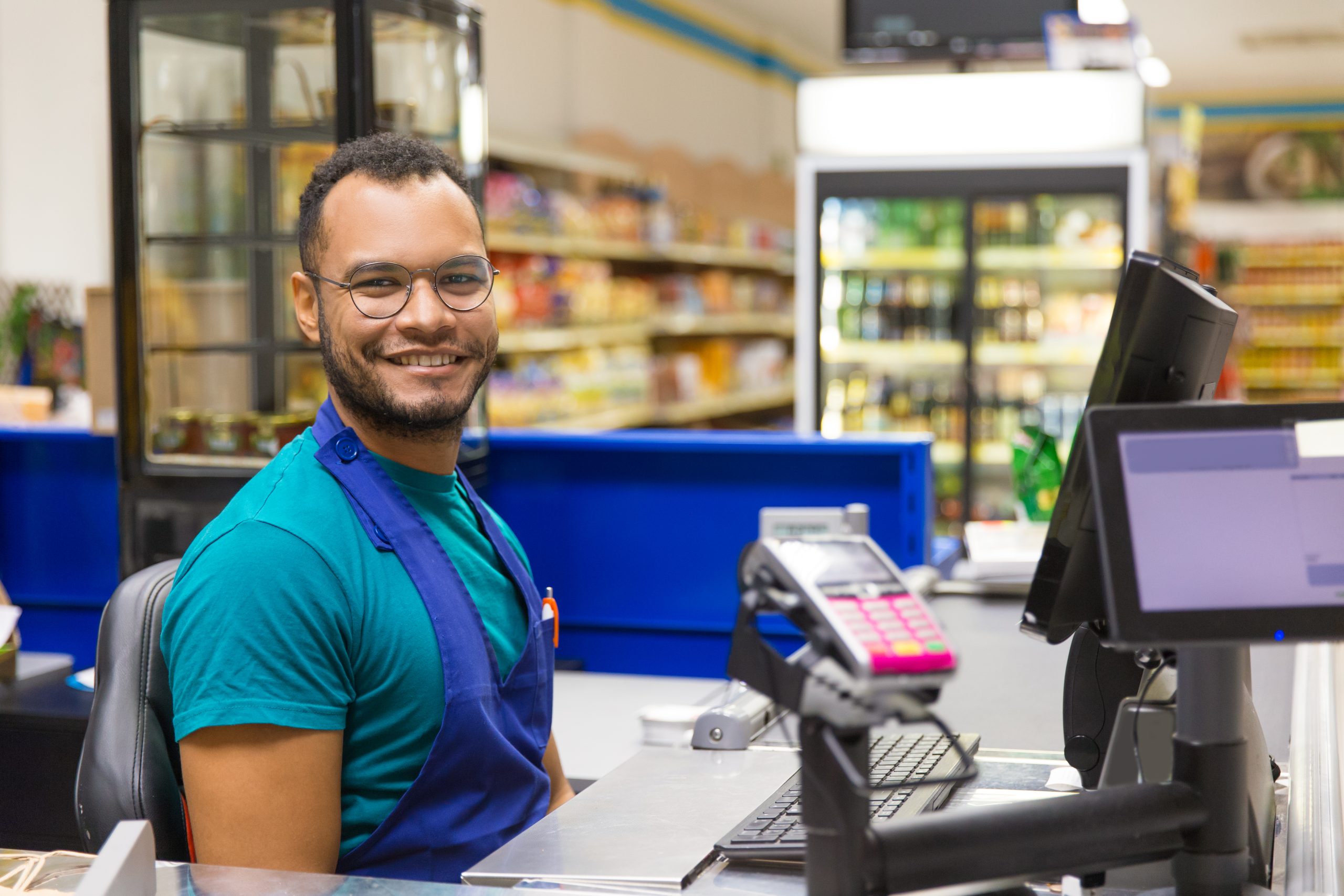 Um homem, que aparenta ser um operador de caixa, está sorrindo para a câmera, sentado em seu posto de trabalho em um supermercado. Ele usa óculos e um avental azul sobre uma camiseta verde-azulada. Ao fundo, percebe-se o corredor da loja com prateleiras de produtos e refrigeradores. A imagem detalha o ambiente de trabalho e, assim, reforça a importância de prevenir erros trabalhistas em supermercados para um ambiente justo.