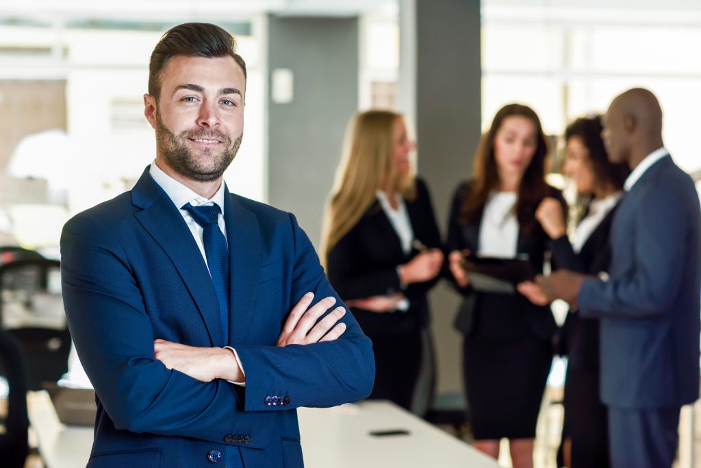 Um jovem empresário sorri para a câmera com os braços cruzados, enquanto colegas trabalham ao fundo. A cena sugere um ambiente corporativo onde a Advocacia Preventiva é fundamental para a segurança jurídica e o bom funcionamento dos negócios.