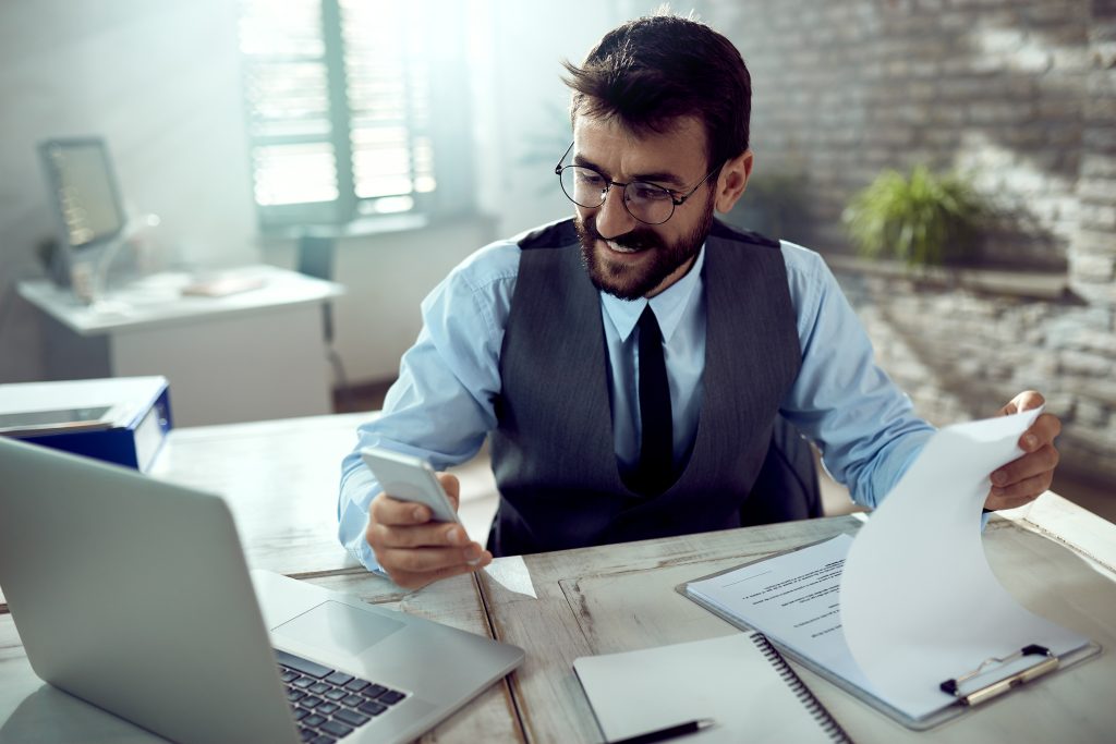 Um jovem empresário com barba e óculos está sorrindo enquanto revisa documentos e usa seu smartphone em um escritório bem iluminado. Ele veste uma camisa social azul, colete e gravata, transmitindo uma imagem de profissionalismo. A cena sugere alguém engajado em tarefas que podem incluir a Análise de impactos legislativos, buscando informações ou confirmando dados importantes.