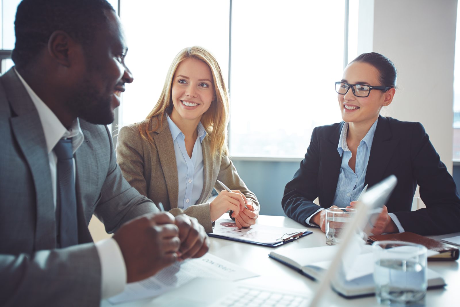 Um grupo de três profissionais, incluindo duas mulheres sorridentes e um homem, reunidos em uma mesa de escritório com um laptop e documentos. A atmosfera colaborativa sugere uma discussão sobre assessoria em processos administrativos.