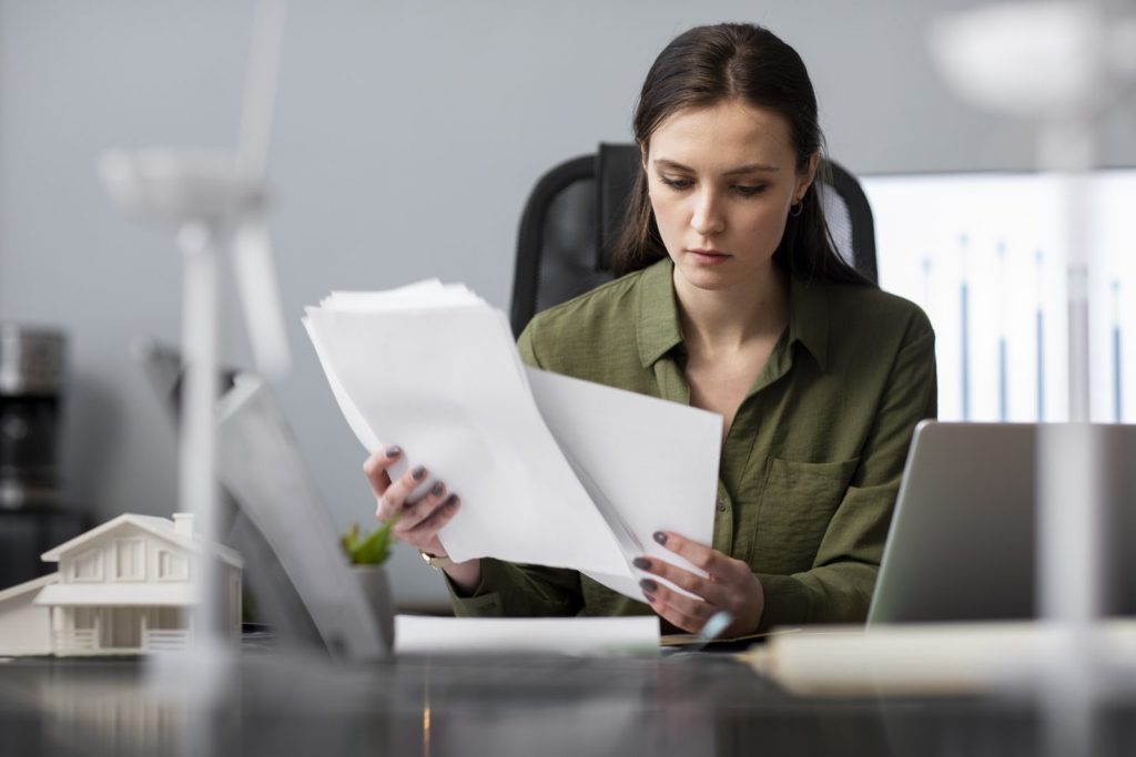 Uma mulher com olhar concentrado e pensativo examina papéis em sua mesa, possivelmente cogitando iniciar uma Auditoria contratual.