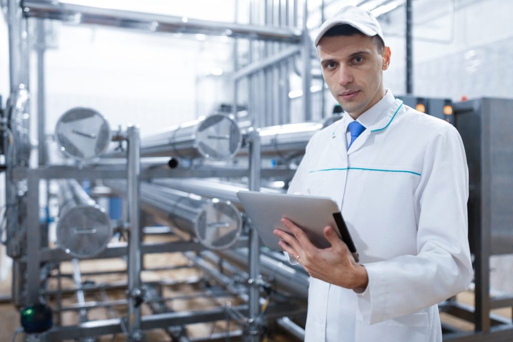 Um inspetor, vestindo um jaleco branco e boné, segura um tablet enquanto inspeciona um maquinário industrial. Seu olhar é direto para a câmera, transmitindo seriedade e competência em um ambiente de controle de produção, o que se alinha bem com a necessidade de assessoria em questões regulatórias para garantir conformidade em processos industriais.
