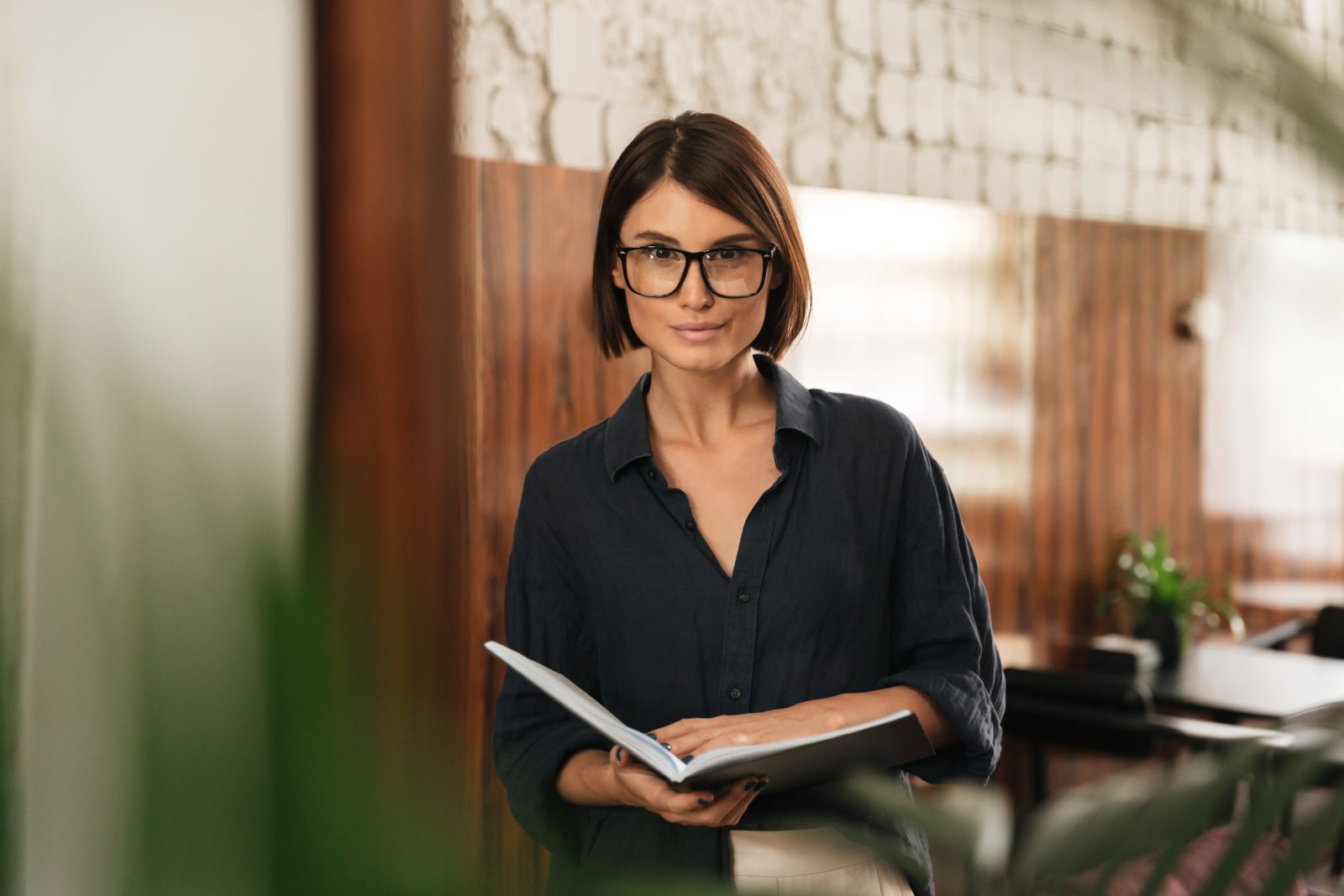 Uma mulher sorridente com cabelos curtos e óculos está segurando um caderno aberto ou documentos. Ela veste uma camisa escura e está em um ambiente interno com paredes de tijolos e elementos de madeira ao fundo, transmitindo uma imagem profissional e atenta, ideal para a área de assessoria em questões regulatórias.
