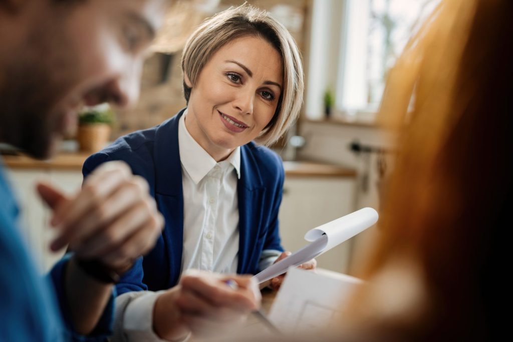 Uma mulher com cabelo curto e loiro, vestindo um blazer azul e camisa branca, sorri enquanto interage com clientes. Ela segura documentos e uma caneta, parecendo guiar uma conversa ou explicar detalhes. A cena sugere uma interação profissional e atenciosa, comum em um processo de auditoria jurídica.