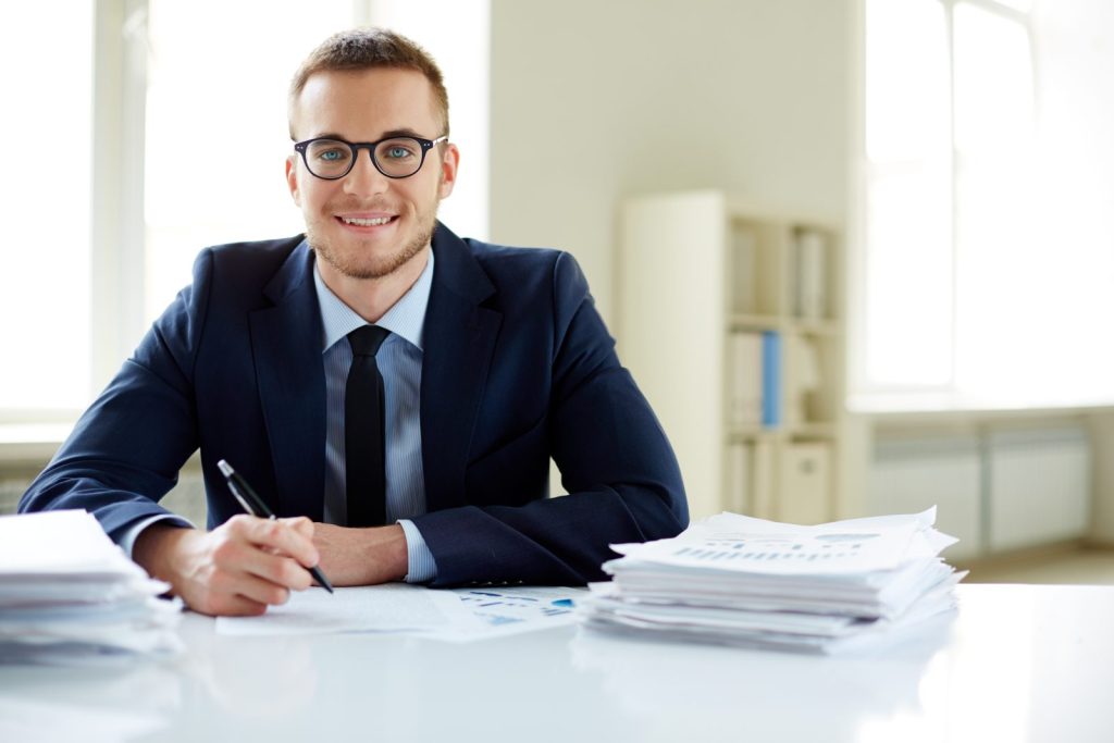 Um executivo jovem e sorridente, usando óculos e um terno azul-marinho, está sentado em sua mesa de escritório. Ele segura uma caneta e tem pilhas de documentos à sua frente, sugerindo um trabalho focado e detalhado, ideal para representar um profissional envolvido em auditoria jurídica.
