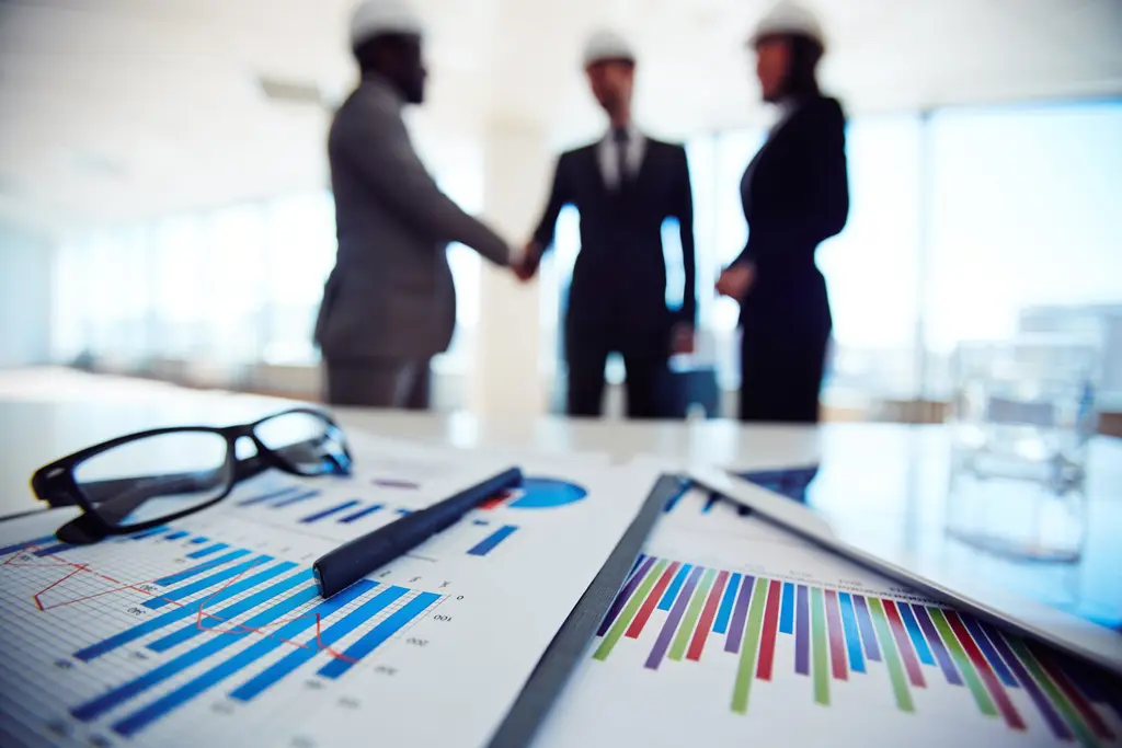 Em primeiro plano, uma mesa de reunião com gráficos financeiros, óculos e uma caneta. Ao fundo, desfocados, três executivos de terno e capacetes de segurança se cumprimentam, possivelmente selando um acordo ou discutindo aspectos de uma auditoria societária.