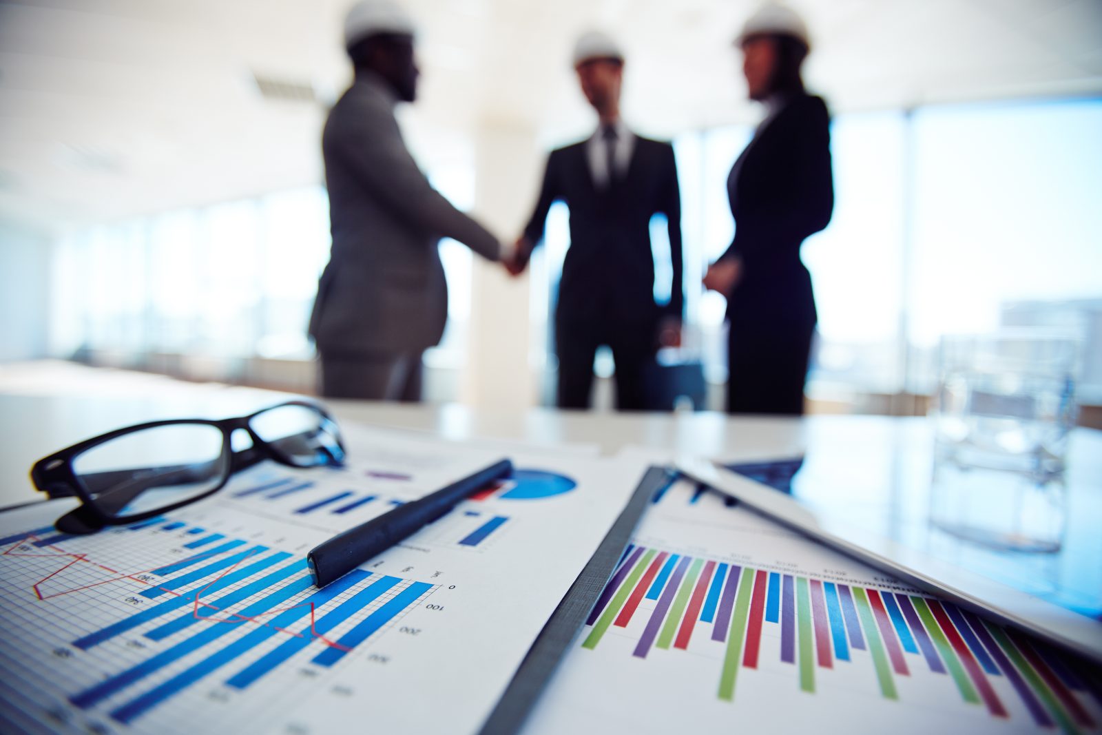 Em primeiro plano, uma mesa de reunião com gráficos financeiros, óculos e uma caneta. Ao fundo, desfocados, três executivos de terno e capacetes de segurança se cumprimentam, possivelmente selando um acordo ou discutindo aspectos de uma auditoria societária.