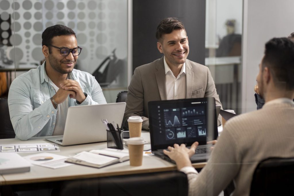 Três homens, um de óculos e camisa casual, outro de blazer, e outro de suéter, sorriem enquanto participam de uma reunião em um escritório moderno. Há laptops abertos com gráficos na tela e copos de café na mesa. A cena sugere um ambiente de negócios colaborativo, onde discussões sobre estratégia e planejamento, como a blindagem patrimonial e societária, podem estar ocorrendo.