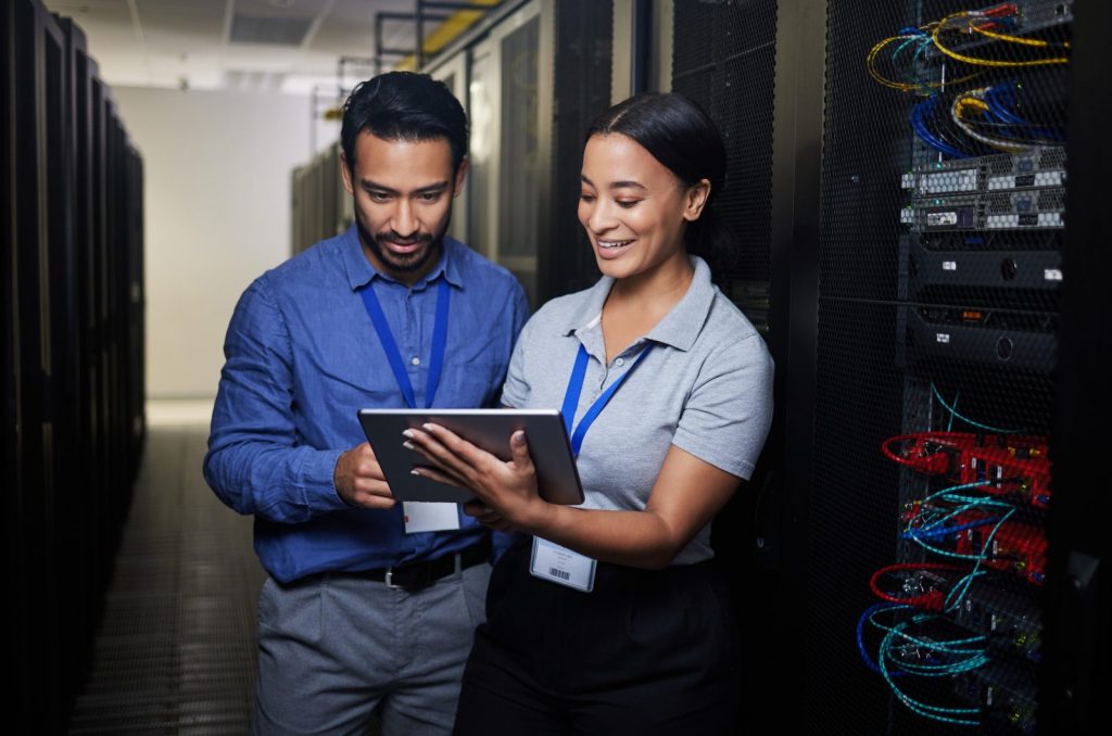 Dois técnicos, um homem e uma mulher, estão inspecionando servidores em um data center. A mulher sorri enquanto ambos olham para um tablet, provavelmente verificando dados ou procedimentos relacionados ao Compliance no setor telecom, garantindo que os sistemas estejam operando conforme as regulamentações.