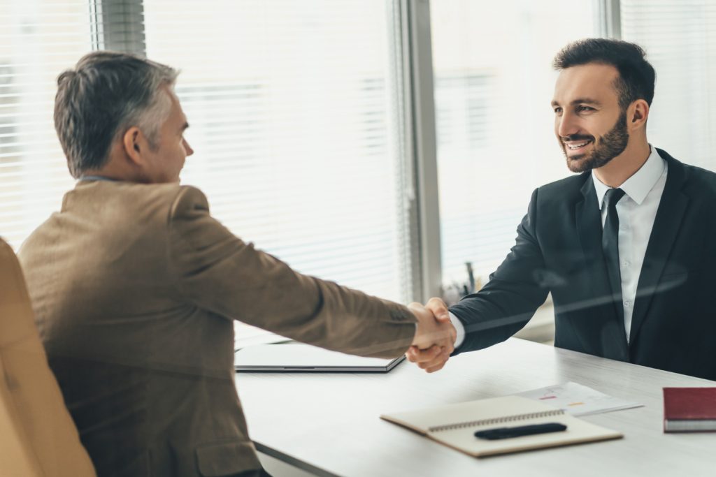 Dois homens de negócios, um mais velho e um mais novo, se cumprimentam com um aperto de mão e sorrisos em um ambiente de escritório. O gesto, feito sobre uma mesa com documentos, sugere que um acordo foi fechado, possivelmente após discutirem as cláusulas contratuais.