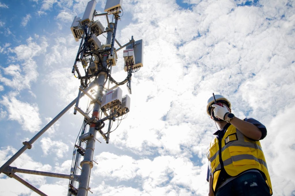 Um engenheiro, usando colete de segurança e capacete, está em pé em uma área aberta com uma torre de telecomunicações ao fundo. Ele segura um rádio comunicador no rosto, conversando com sua equipe. A imagem transmite a ideia de comunicação e trabalho no campo, o que é fundamental para o Compliance no setor telecom, assegurando que a infraestrutura e as operações estejam de acordo com as normas.