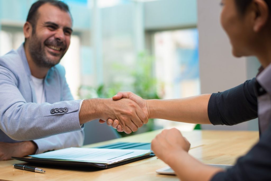 Duas pessoas sorridentes apertam as mãos sobre uma mesa de escritório, onde documentos estão visíveis, confirmando a formalização de Contratos com fornecedores.