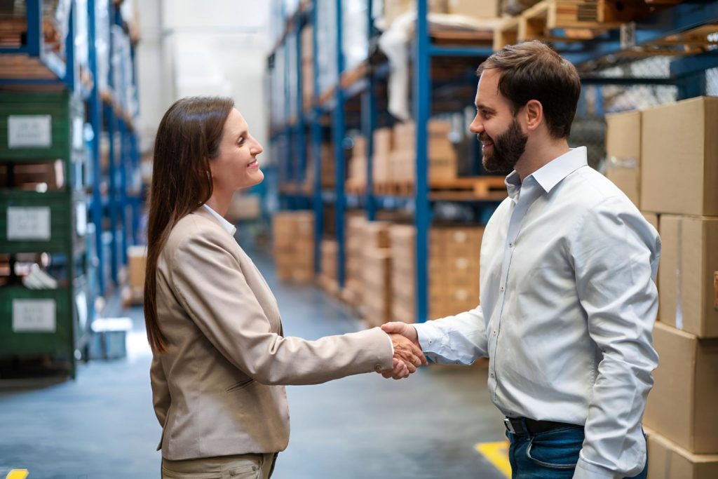 Um homem e uma mulher se cumprimentam com um aperto de mão em um armazém, em meio a prateleiras e caixas de papelão, sinalizando a finalização de Contratos com fornecedores.