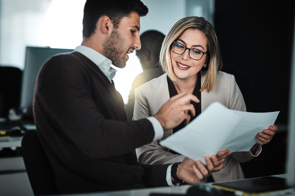 Um homem e uma mulher, ambos sorridentes, estão sentados em uma mesa, revisando um documento. O homem aponta para o papel enquanto a mulher o segura, sugerindo que estão em uma colaboração para analisar contratos comerciais.