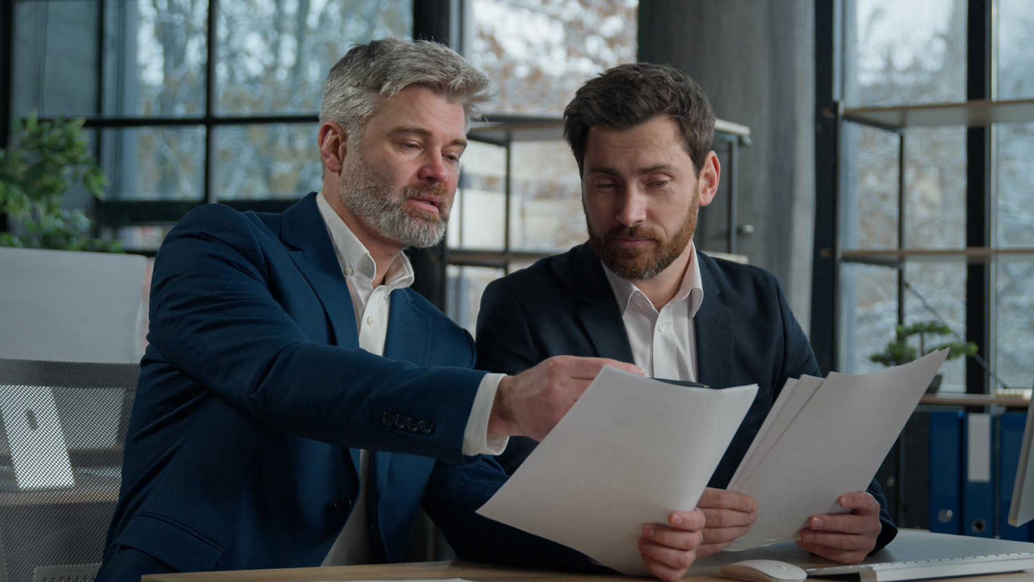 Dois homens de negócios, um mais velho com barba grisalha e um mais jovem, estão sentados em uma mesa, revisando uma pilha de documentos. O mais velho aponta para um dos papéis, transmitindo seriedade e foco na análise dos contratos empresariais.
