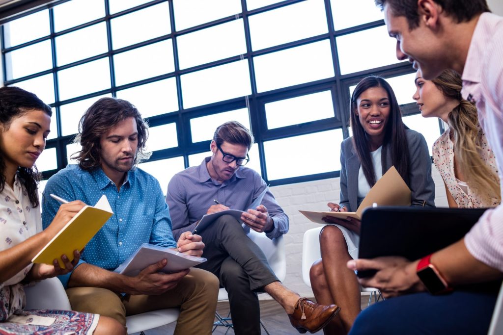 Um grupo de seis colegas de trabalho em uma reunião, sentados perto de uma grande janela, interagindo e fazendo anotações em pastas e cadernos. Eles demonstram colaboração e discussão de documentos, sugerindo a implementação de uma cultura jurídica preventiva na empresa.