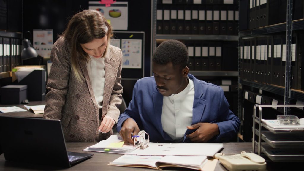 Homem e mulher de negócios examinando minuciosamente documentos em uma mesa com muitas pastas arquivadas.