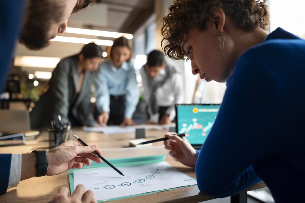 Imagem que foca em dois profissionais de negócios, um homem e uma mulher, analisando e apontando para um diagrama de fluxo em papel em uma mesa, com outros colegas trabalhando no fundo. O olhar sério e a análise de gráficos financeiros no laptop ao lado sugerem a discussão de riscos e estratégias para evitar uma possível falência.