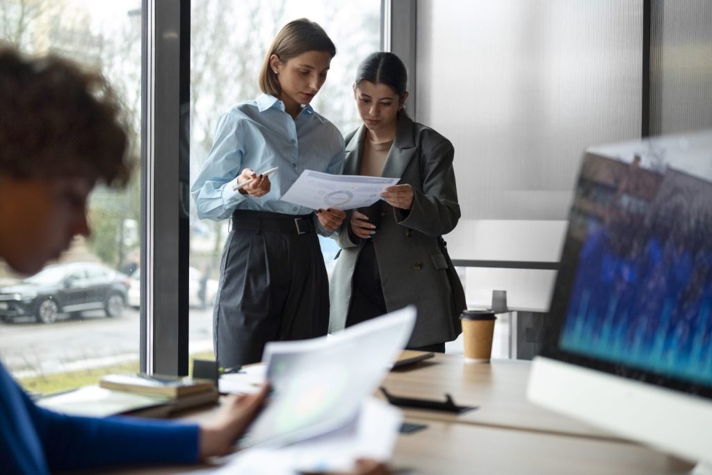 Duas mulheres em pé analisam documentos, colaborando na gestão de contratos no escritório. 
