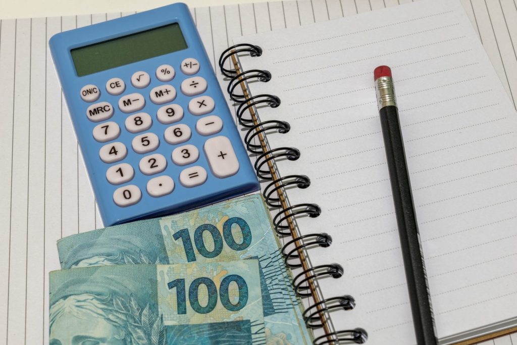 Detalhe de notas de 100 Reais, uma calculadora azul e um caderno espiral com um lápis, simbolizando a gestão de perdas tributárias e o controle financeiro.