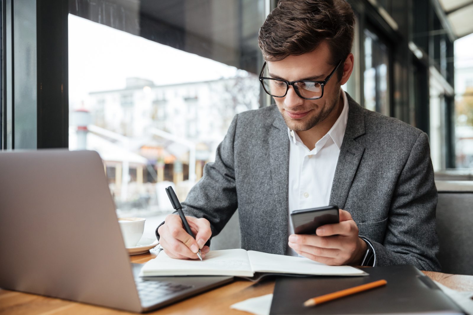 Empresário sorridente de óculos, sentado em um café, usando o smartphone enquanto anota em um caderno ao lado de seu laptop. A cena ilustra a gestão de riscos fiscais em um ambiente de trabalho híbrido.