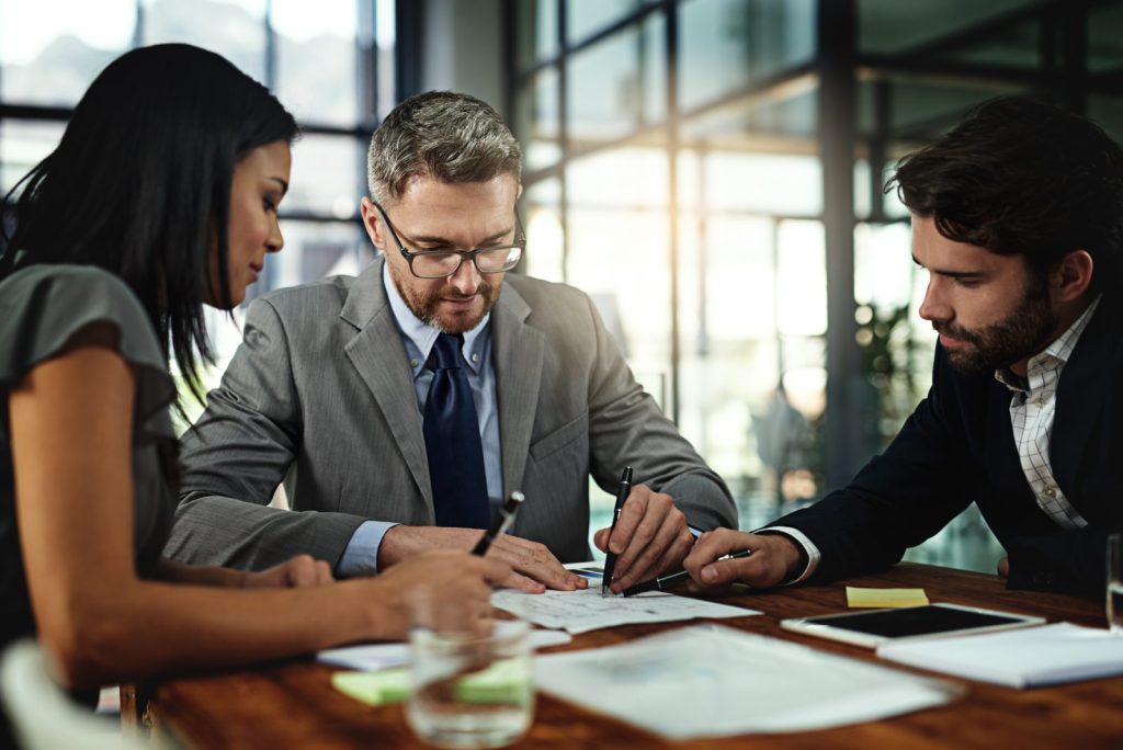 Advogado de terno cinza e óculos analisa contrato na mesa com seus clientes em consultoria jurídica empresarial.