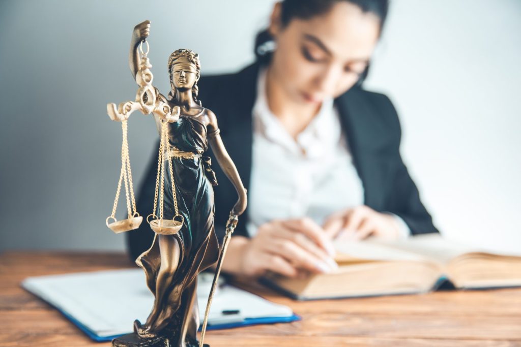 Estátua da justiça em primeiro plano e advogada estudando ao fundo, simbolizando a representação judicial empresarial.