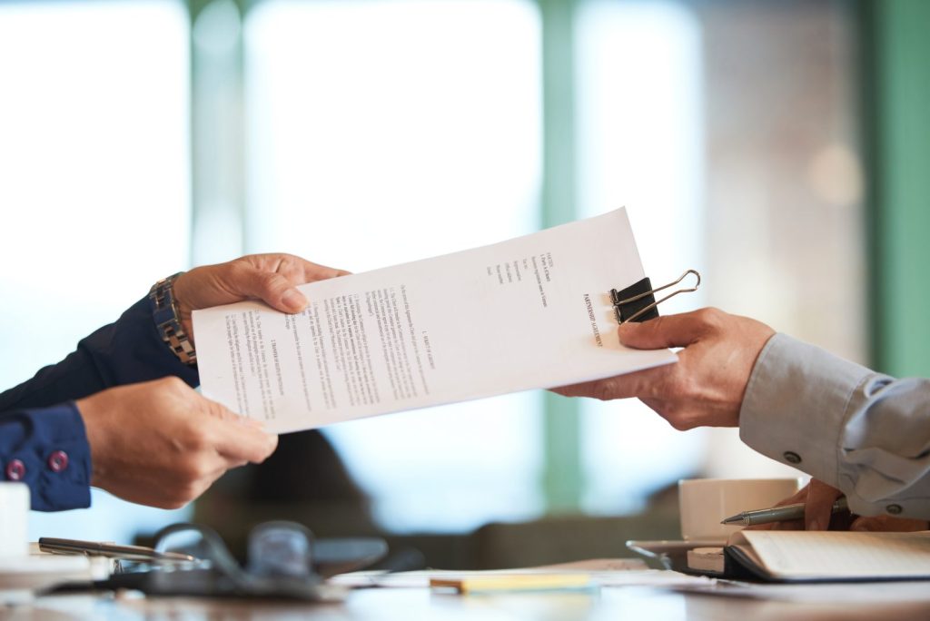 Mãos trocando documentos em reunião, ilustrando a formalização de contratos trabalhistas entre empresa e colaborador.